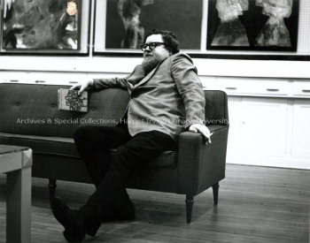 Alden Nowlan sitting on a sofa in the Art Centre, with a book in one hand and a cigarette in the other hand. UA PR; Series 2, Sub-series 4, File 1021, Item 1